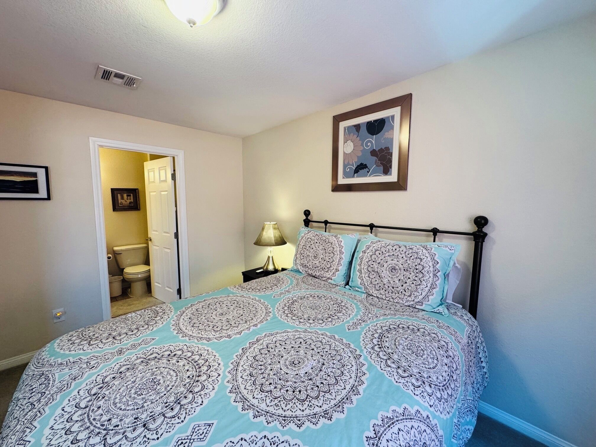 Un dormitorio con cama, mesita de noche, lámpara, un cuadro en la pared y una puerta abierta que da al baño.