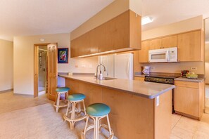 A modern kitchen with wooden cabinets, a stainless steel refrigerator, a microwave, and a built-in stove. There are three bar stools with teal cushions. - Maui Sunset Vacation Rentals A-306 (Kihei)