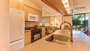 A modern kitchen with a refrigerator, microwave, stove, and sink. There is a dining area with a table and chairs, and a view of the outdoors through large windows. - Maui Sunset Vacation Rentals A-306 (Kihei)