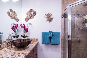 A bathroom with a marble countertop, a large decorative bowl, two potted orchids, and a glass shower enclosure.