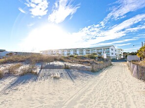 Een hotel aan het strand, met een zandpad naar de ingang, omgeven door begroeiing en een heldere lucht.