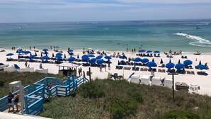 Ein Strand mit vielen blauen Sonnenschirmen, einem sandigen Ufer und einem wolkenlosen, blauen Himmel.