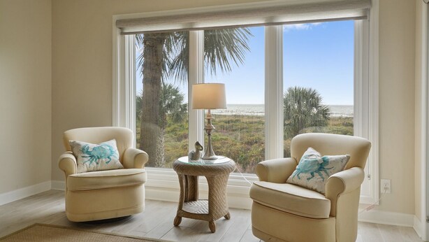 A room with two armchairs, a small round table with a lamp, and a large window offering a view of the beach and palm trees. - Shorewood 202 - Direct Oceanfront Beautiful Condo!! (Hilton Head Island)