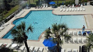 A large rectangular swimming pool surrounded by lounge chairs and umbrellas, with palm trees and a building in the background. - Shorewood 202 - Direct Oceanfront Beautiful Condo!! (Hilton Head Island)