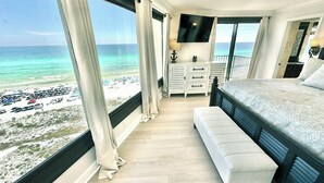 A bedroom with a large window overlooking a beach, a bed, a bench, and a TV. - Large corner unit with spacious balconies! 4106 BS1 (Miramar Beach)