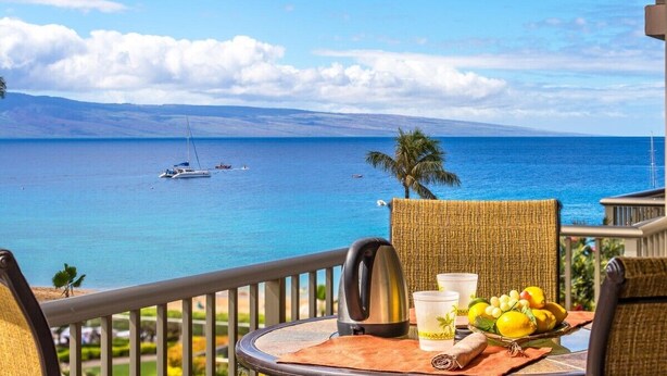 A balcony with a table set for two, overlooking a clear blue sea with boats, palm trees, and a distant mountain range. - Whaler 523 - Amazing Oceanfront on Kaanapali Beach (Lahaina)