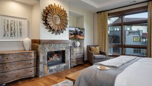 Un dormitorio moderno con chimenea de piedra, un espejo grande y vistas a una zona residencial a través de un ventanal amplio.
