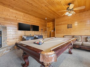 A pool table with billiard balls, two foosball tables, a flat-screen TV, and a ceiling fan in a wooden room.