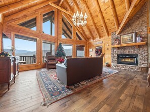 A spacious living room with a fireplace, wooden beams, large windows, and a cozy fireplace mantel. - Cabin w/Amazing Views, Large Game Room, Hot Tub (Sevierville)