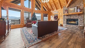 A spacious living room with a fireplace, wooden beams, large windows, and a cozy fireplace mantel. - Cabin w/Amazing Views, Large Game Room, Hot Tub (Sevierville)