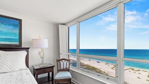 A bedroom with a large window overlooking a beach, a bed with a quilted cover, a nightstand with a lamp, and a chair.