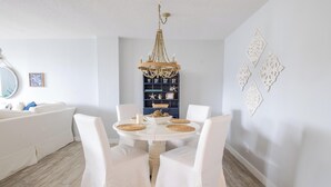 A dining area with a round table, white chairs, a chandelier, and a wall with decorative elements. - Direct Beach Access - Poolside Coastal Condo: Sea Gem (Palm Beach Shores)