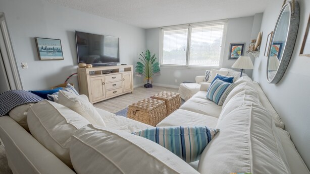 A modern living room with a large white sofa, a flat-screen TV, a wooden cabinet, and a potted plant. - Direct Beach Access - Poolside Coastal Condo: Sea Gem (Palm Beach Shores)