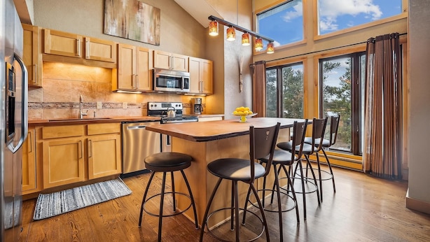 A modern kitchen with wooden cabinets, a stainless steel refrigerator, a microwave, and a stove. There is a dining area with a table and four stools. Large windows provide natural light and a view of the outdoors. - 4 O'Clock A16: Ski-In Condo, Gas Fireplace, 1 Block to Snowflake Lift. (Breckenridge)