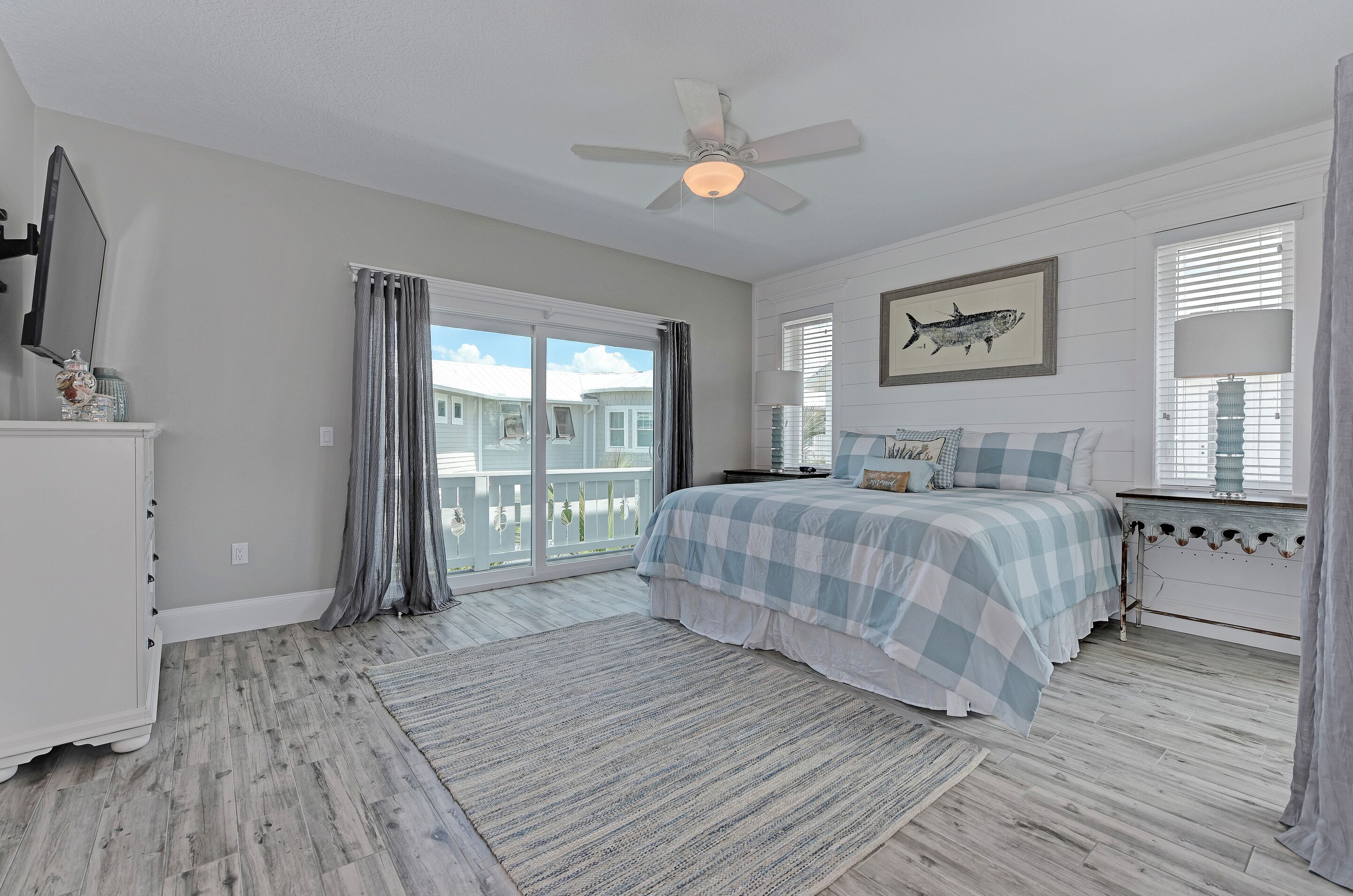 A bedroom with a large bed, a ceiling fan, a window with curtains, and a view of a house outside.