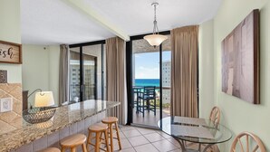 A modern kitchen with a granite countertop, wooden stools, a glass dining table, and a view of the ocean through large sliding doors.