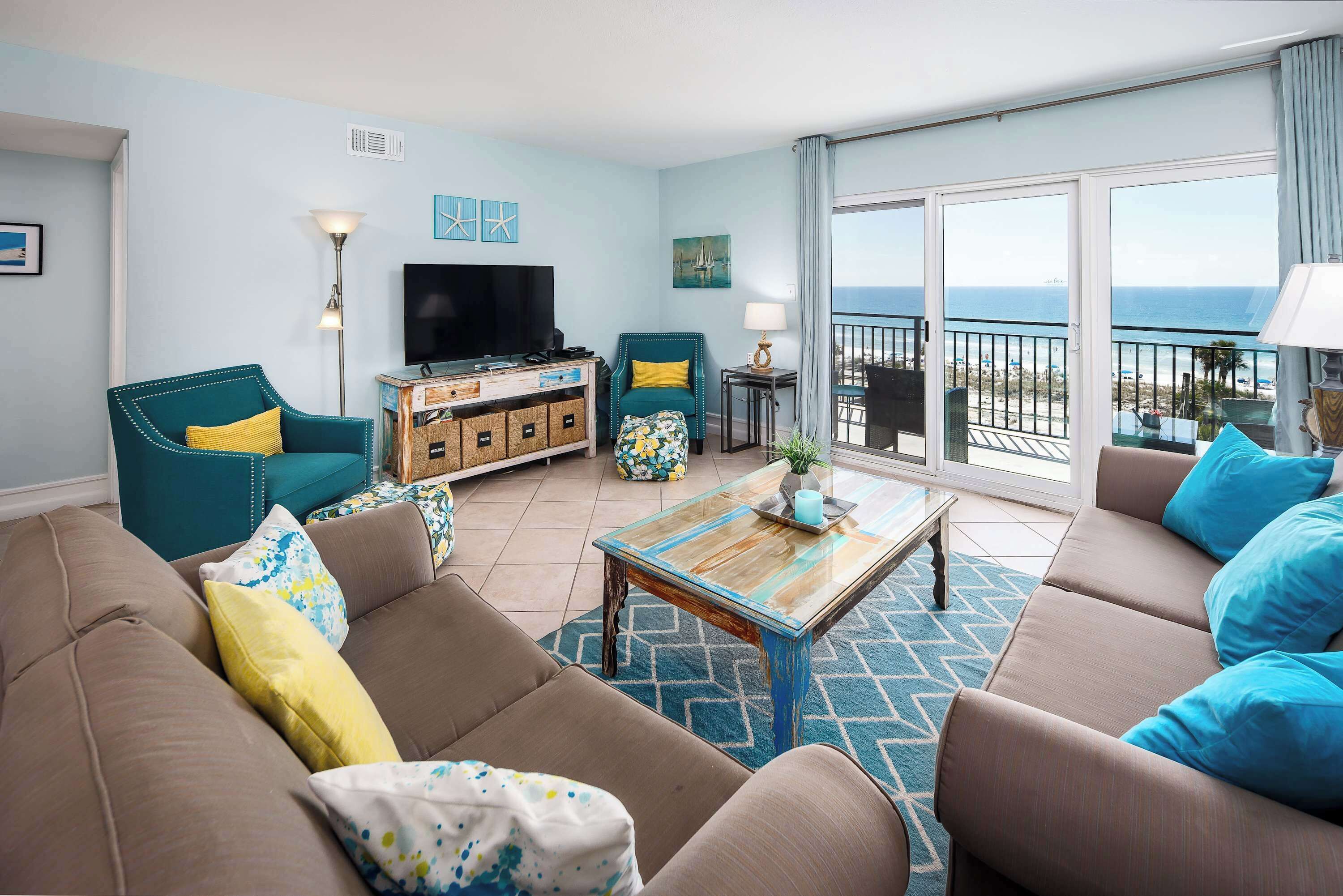 A living room with a sofa, armchairs, a coffee table, and a TV. There is a balcony with a view of the ocean.
