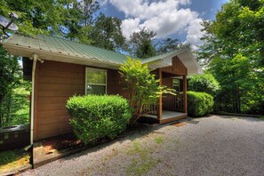 A wooden cabin with a porch, surrounded by trees and a gravel driveway.