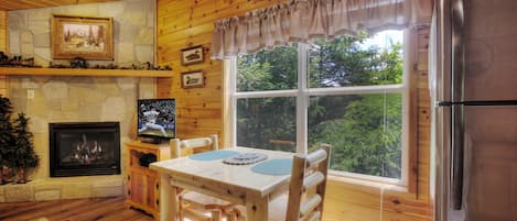 A wooden cabin interior with a stone fireplace, a dining table with chairs, a large window, and a refrigerator.