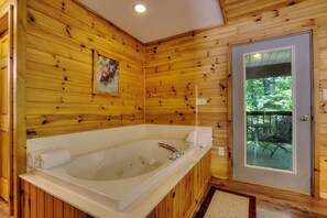 A wooden cabin bathroom with a large bathtub, wooden walls, a door with a view of the outdoors, and a towel on the tub.