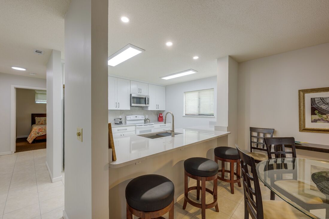 A modern kitchen with white cabinets, a stainless steel microwave, and a glass dining table with four stools.