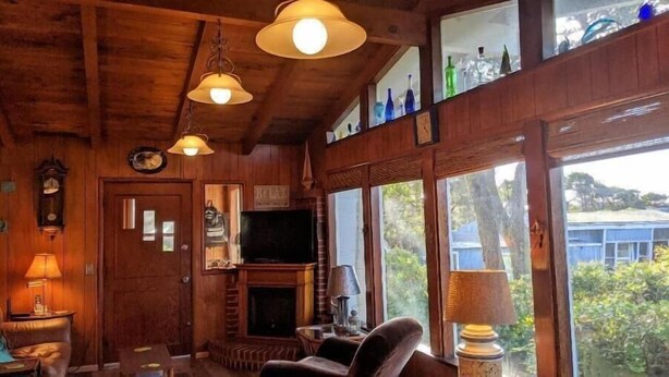 A wooden interior with a fireplace, television, and large windows overlooking greenery. - On Beach Time – Vintage Cottage Near the Surf (Florence)