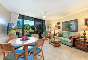 A spacious living area with a round dining table, wicker furniture, a sofa with tropical prints, and a large window offering a view of greenery.
