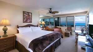 Une chambre d’hôtel équipée d’un lit, d’un bureau, d’une chaise, d’une lampe, d’un ventilateur de plafond et d’une grande fenêtre donnant sur l’océan.