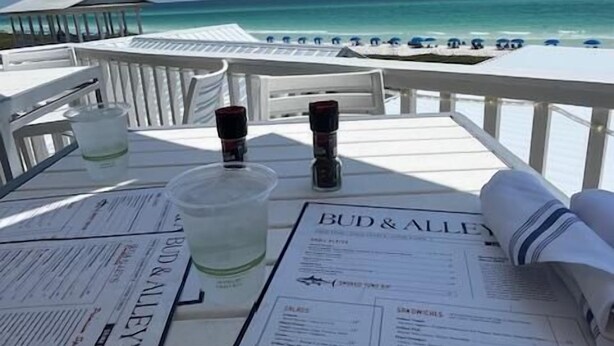A table with a menu, glasses, and a bottle, overlooking a beach with umbrellas. - Bay View,  Villa Golf Cart, Private Deck, Blue Bayou 270 Fairways (Miramar Beach)