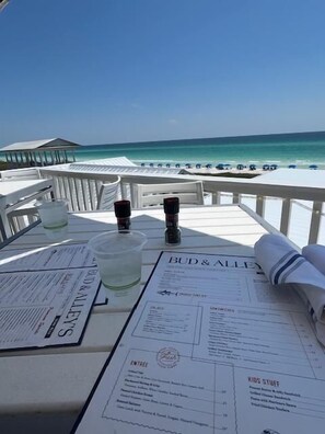 Een tafel met een menukaart, glazen en een fles, met uitzicht op een strand met parasols.