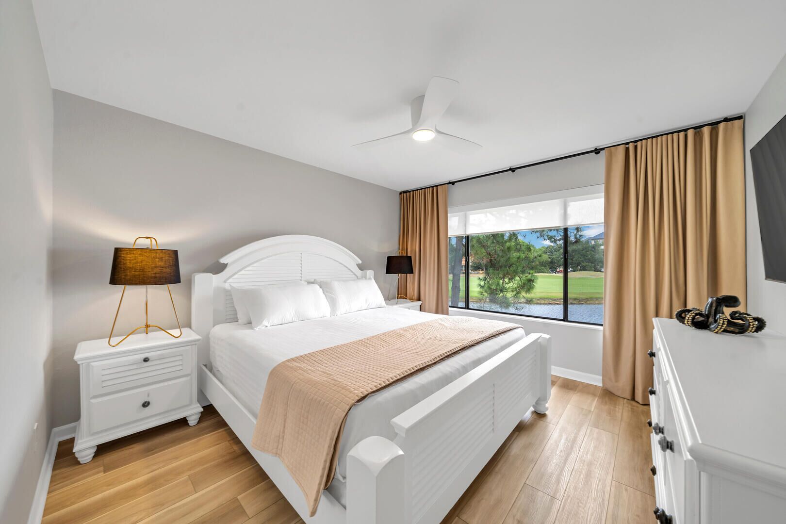 A modern bedroom with a large bed, bedside tables, a ceiling fan, and a view of a golf course through the window.