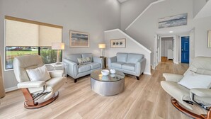 A spacious living room with a high ceiling, a ceiling fan, and a staircase leading to an upper level. There are two light-colored sofas, a glass coffee table, and a rocking chair. - Bay View,  Villa Golf Cart, Private Deck, Blue Bayou 270 Fairways (Miramar Beach)