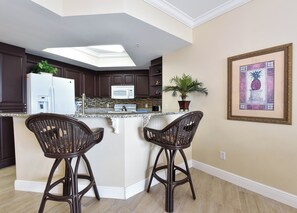 A kitchen with dark wood cabinets, a white refrigerator, a microwave, and a granite countertop. There are two wicker bar stools and a potted plant on the counter.