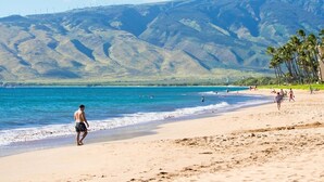Une plage où l’on voit des gens se promener et nager, des palmiers et des montagnes en arrière-plan.