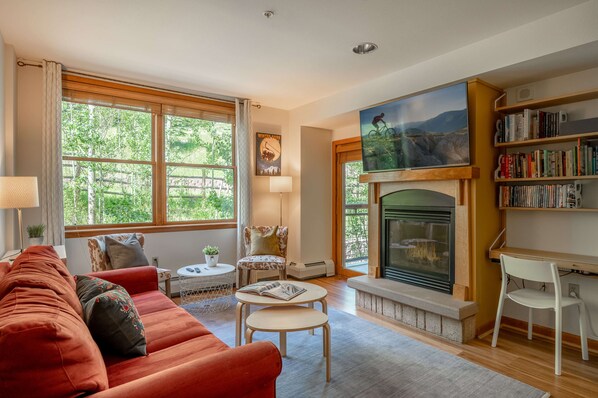 A cozy living room with a fireplace, a bookshelf, and a large window with a view of greenery. - ☀ Sunny Base Area Condo | Zephyr Mountain Lodge 1303 | Convenient Location | Direct Slope Views! (Winter Park)