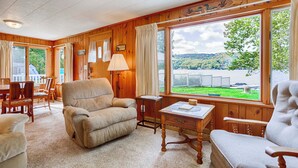 Une chambre confortable, aménagée dans le style d’un chalet, avec une grande fenêtre donnant sur le lac, des murs en bois et un coin salon agréable avec un fauteuil et une table.