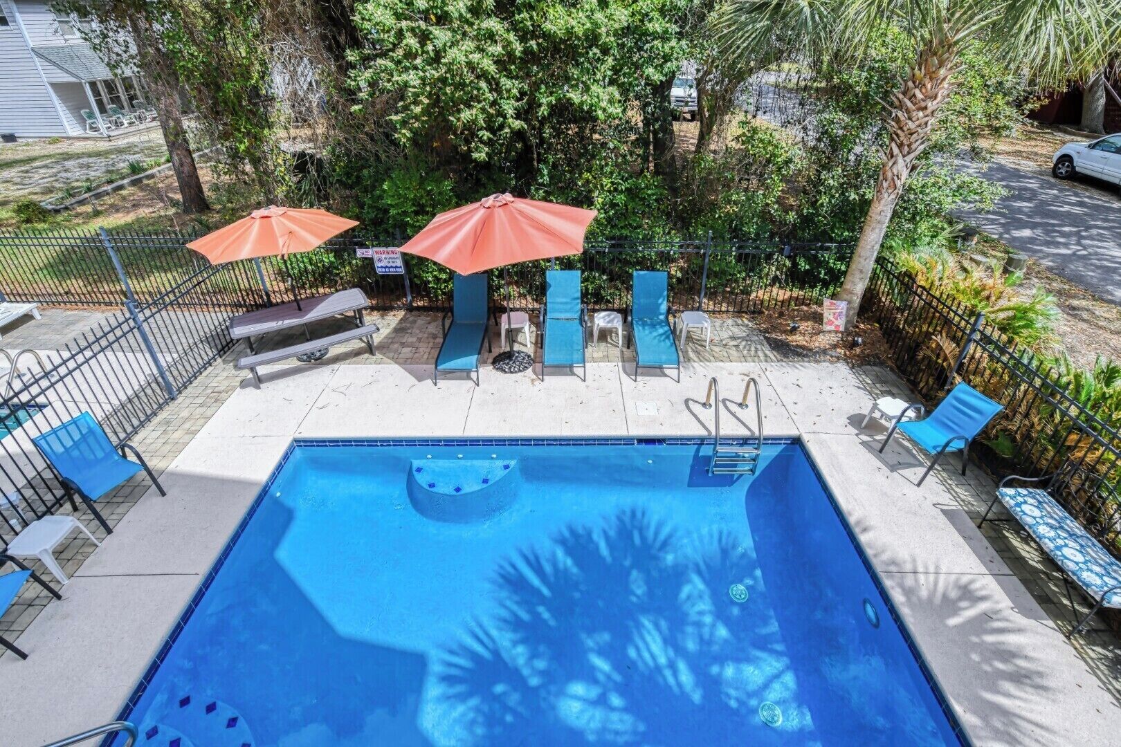 A rectangular swimming pool with a diving board, surrounded by a fence, and shaded by umbrellas.