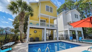 A two-story building with a pool, outdoor seating, and a palm tree. - A Wave from it All - Five Bedroom, Four Bath Beach House (Surfside Beach)