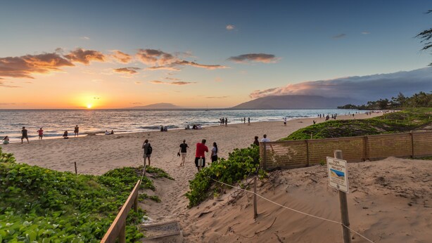 Una playa con gente caminando y sentada, un atardecer al fondo y un letrero que indica la restauración de las dunas.