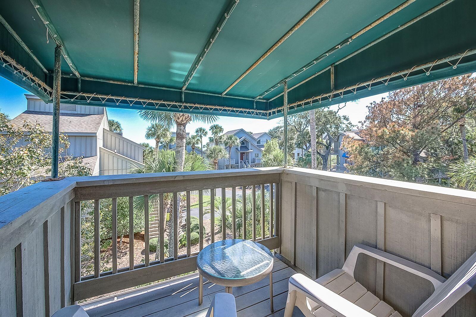 Un balcon abrité, doté d’une table et de chaises en verre, qui offre une vue sur un quartier résidentiel avec ses maisons et ses palmiers.