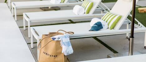 Una hilera de sillas reclinables blancas con almohadillas a rayas verdes y blancas, un bolso marrón con la etiqueta 'AVANTSTAY' en el suelo y un poste de metal al frente.