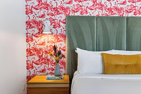 Una camera da letto con una testiera imbottita verde, un letto bianco con un cuscino color senape, un comodino con una lampada e una parete con motivo a fenicottero.