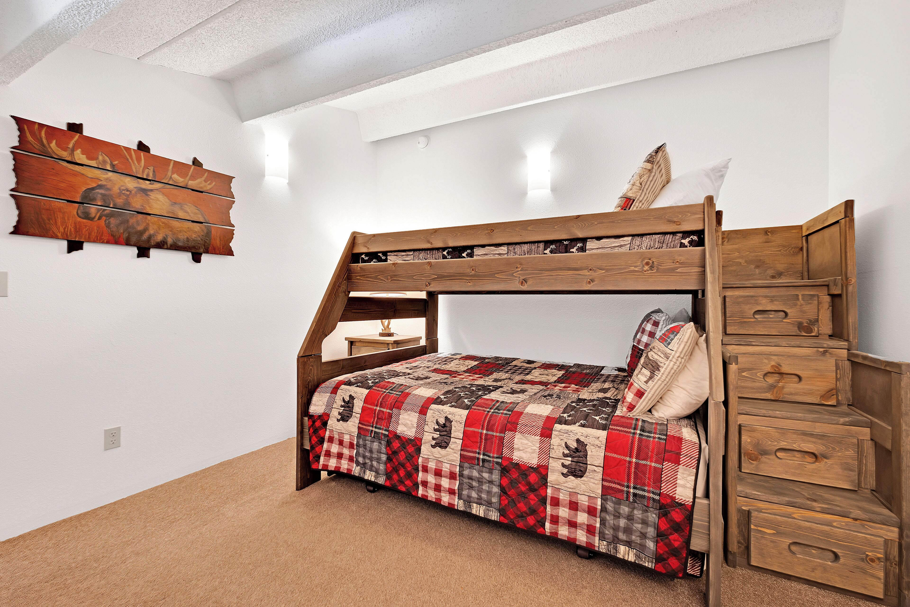 Una cama litera con colcha a cuadros, cajones de madera y un cuadro de un alce en la pared.