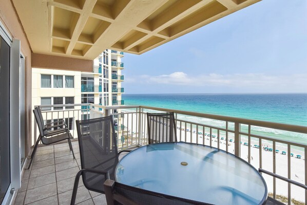 A balcony with a glass table and chairs, overlooking a beach with a clear view of the ocean and a building in the background.