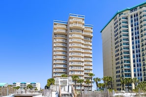 A multi-story hotel with balconies, located near a beach with palm trees.