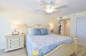 A bedroom with a bed, nightstands, a ceiling fan, and a door leading to another room.