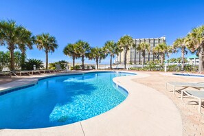 A swimming pool with a curved edge, surrounded by palm trees and lounge chairs, with a hotel building in the background. - Beachfront w/panoramic views, balcony, & beach access! (Destin)