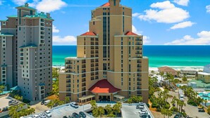 Un hôtel à plusieurs étages, avec un bâtiment central doté d’un toit rouge, entouré d’un parking et de palmiers, et offrant une vue sur la plage.