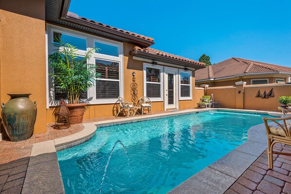 A swimming pool with a tiled deck, surrounded by a house with a tiled roof and a yellow wall.