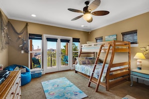 A cozy bedroom with a bunk bed, a ceiling fan, a window with a view, and a small desk with a lamp.